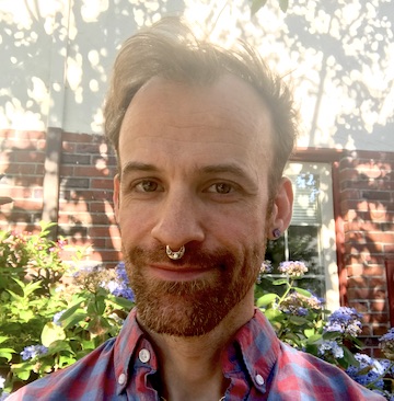 Photo of Sasha Strong standing in front of an apartment building with light and shade from a tree and flowery bushes in the background.