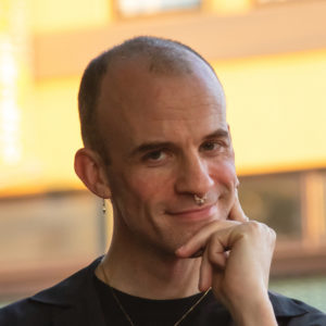 Headshot of Sasha Strong. Sasha is a white person with a shaved head, earrings, a gold nose ring, a necklace, and black dress. Their hand is lifted to their face touch their chin and cheek. They are smiling and looking at the camera. The background is a sunlit, out-of-focus urban scene.